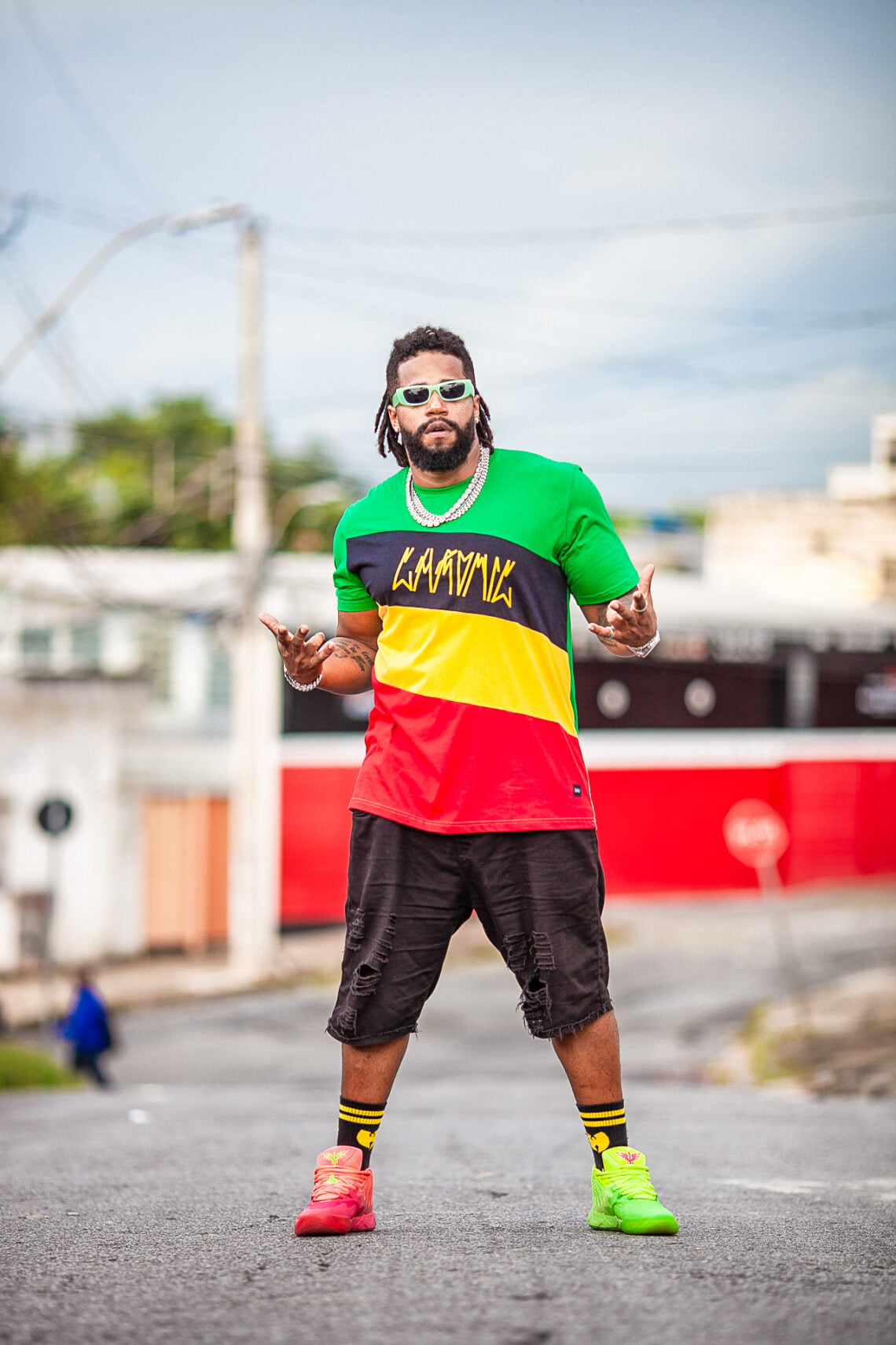 foto do artista Adam Araújo em uma rua fazendo uma pose para o fotógrafo. O artista é um homem negro, com os cabelos em dreads, blusa nas cores verde amarelo preto e vermelho e bermuda preta. Ele usa óculos escuros e um colar.