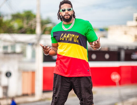 foto do artista Adam Araújo em uma rua fazendo uma pose para o fotógrafo. O artista é um homem negro, com os cabelos em dreads, blusa nas cores verde amarelo preto e vermelho e bermuda preta. Ele usa óculos escuros e um colar.